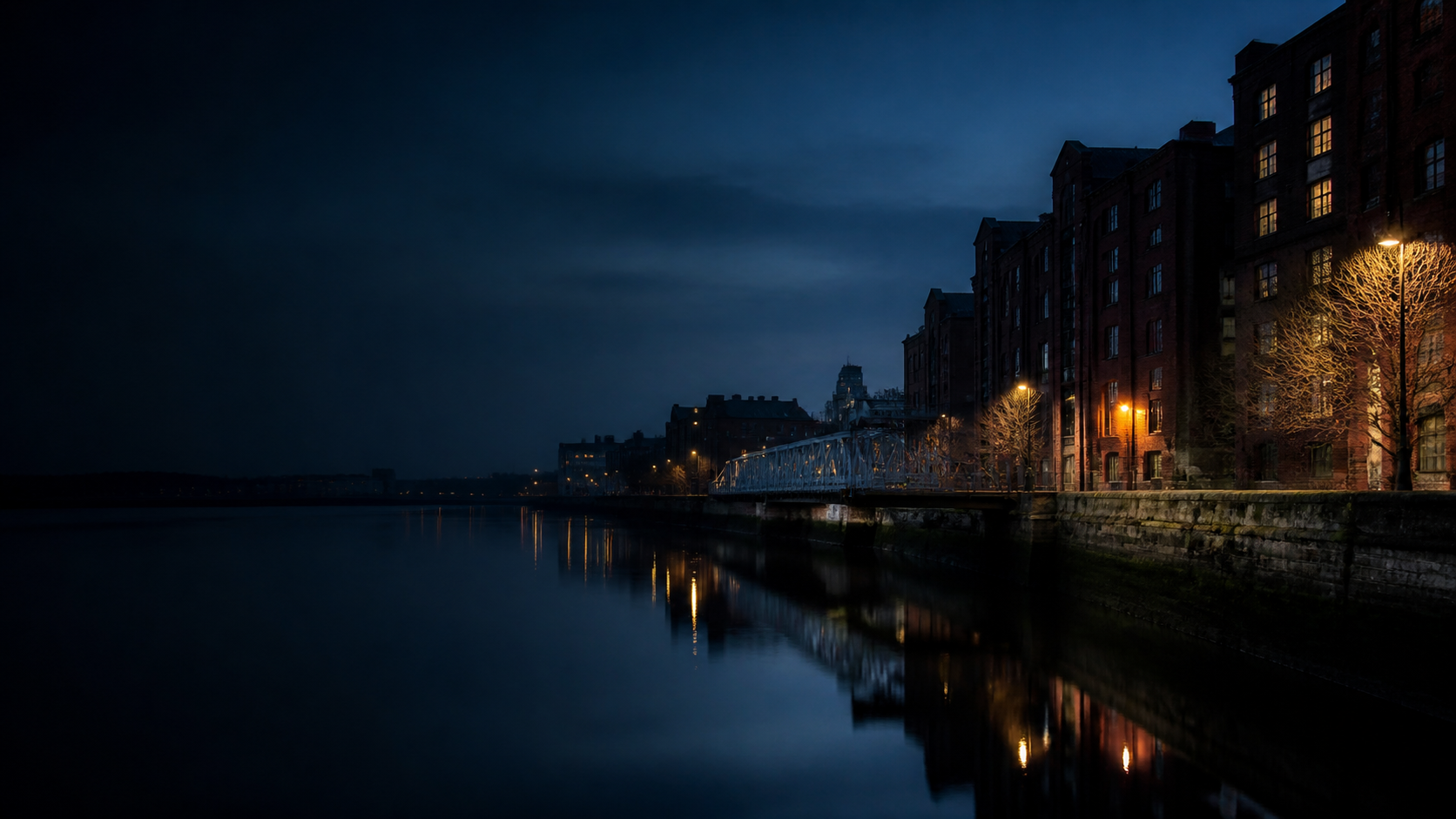 An atmospheric northern riverside at dusk, with dark brick buildings and a steel bridge reflected in the water.